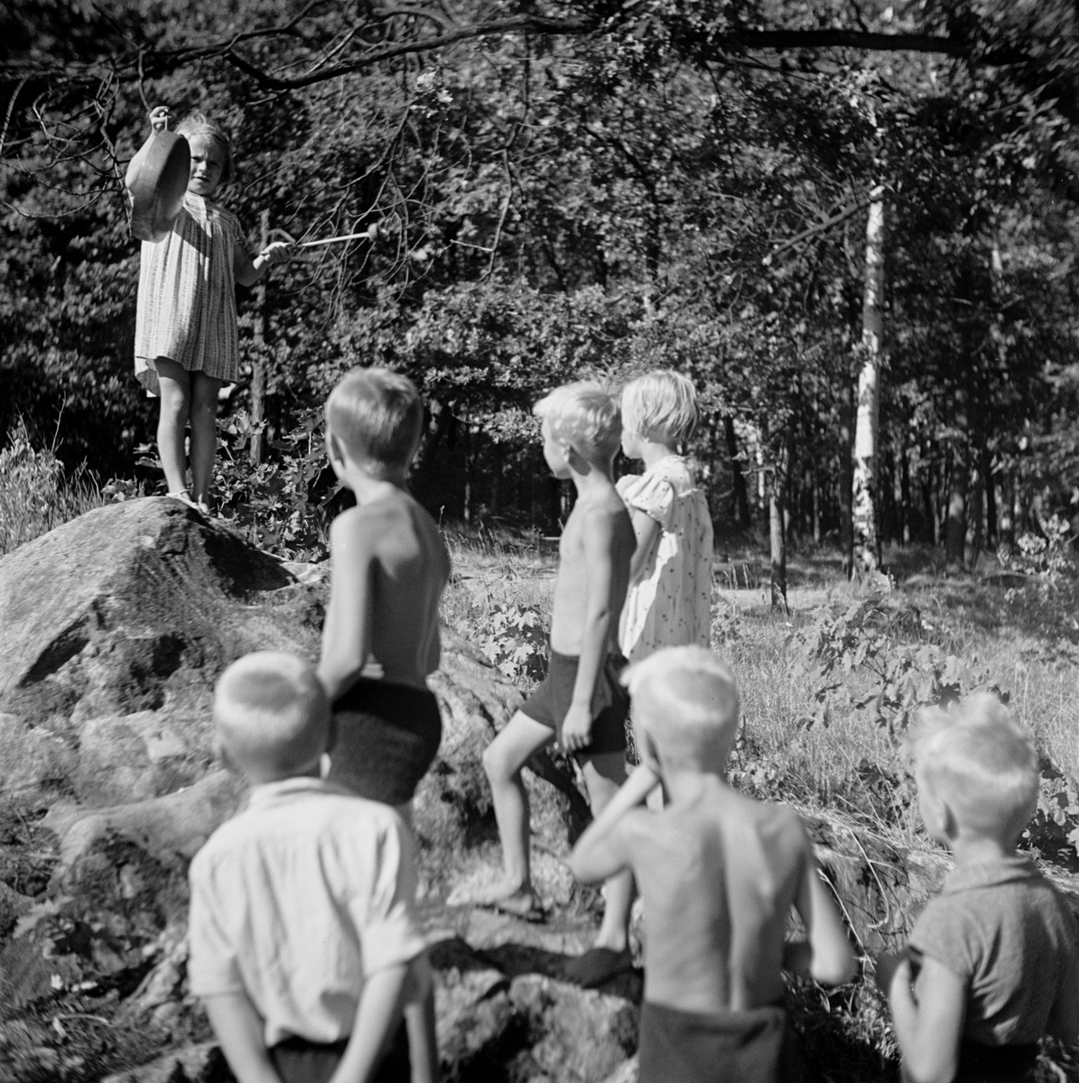Tyttö gongeineen (kattila) kivellä edessään muita lapsia, gongilla ilmoitettiin aterioista