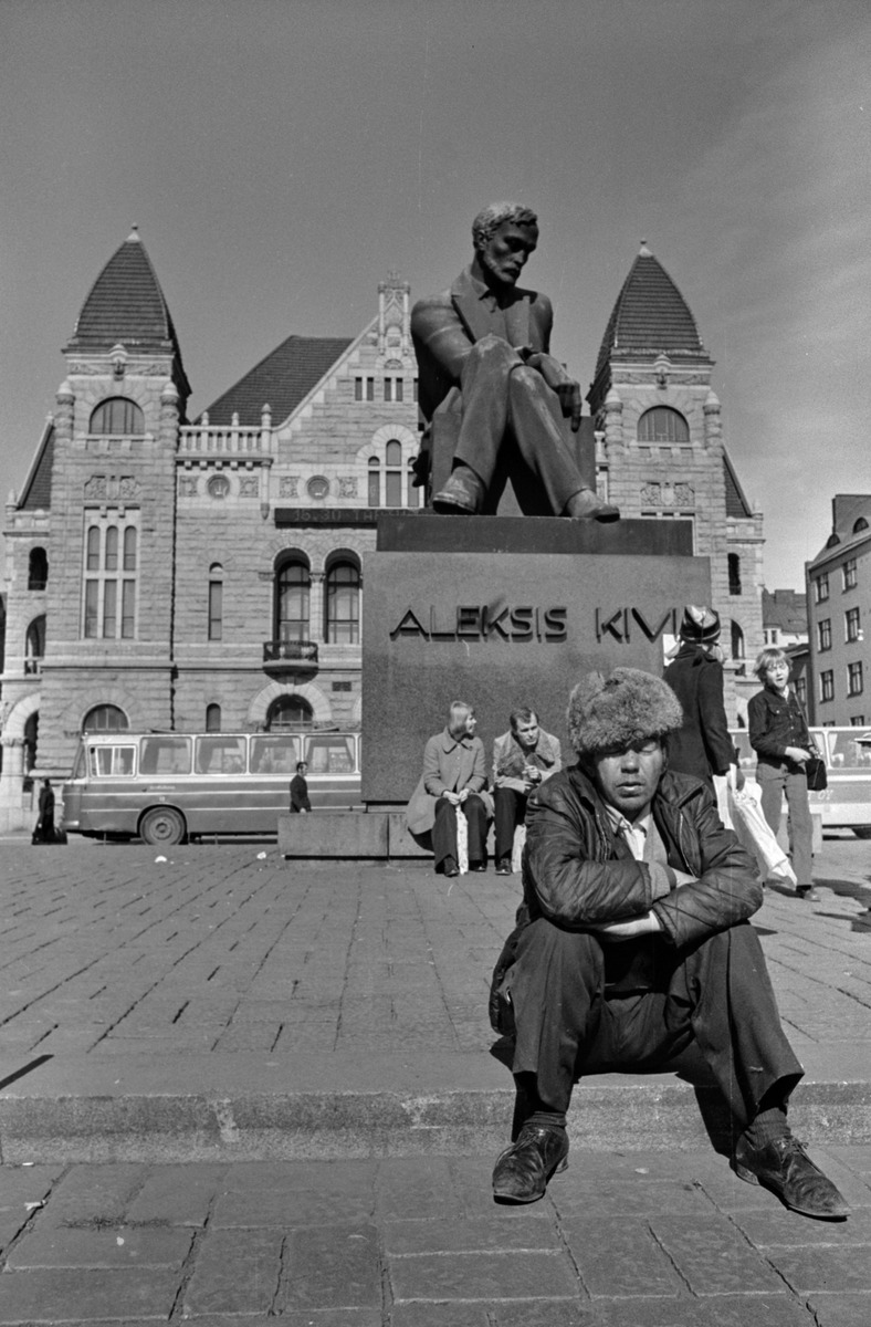 Likaisiin vaatteisiin pukeutunut, karvalakkipäinen mies istuu maassa Rautatientorilla Aleksis Kiven patsaan edessä