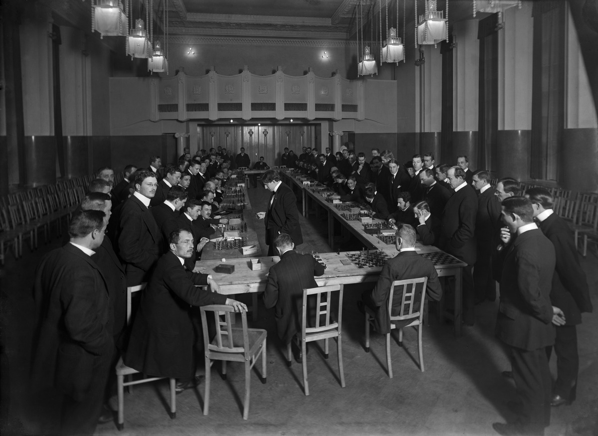 Yhdysvaltalainen mestari Frank James Marshall (1877-1944) pelaa simultaanishakkia Helsingin Shakkiklubin ja Akateemisen Shakkiklubin jäseniä vastaan Uuden Ylioppilastalon (arkkitehdit Armas Lindgren, Wivi Lönn 1910) juhlasalissa.