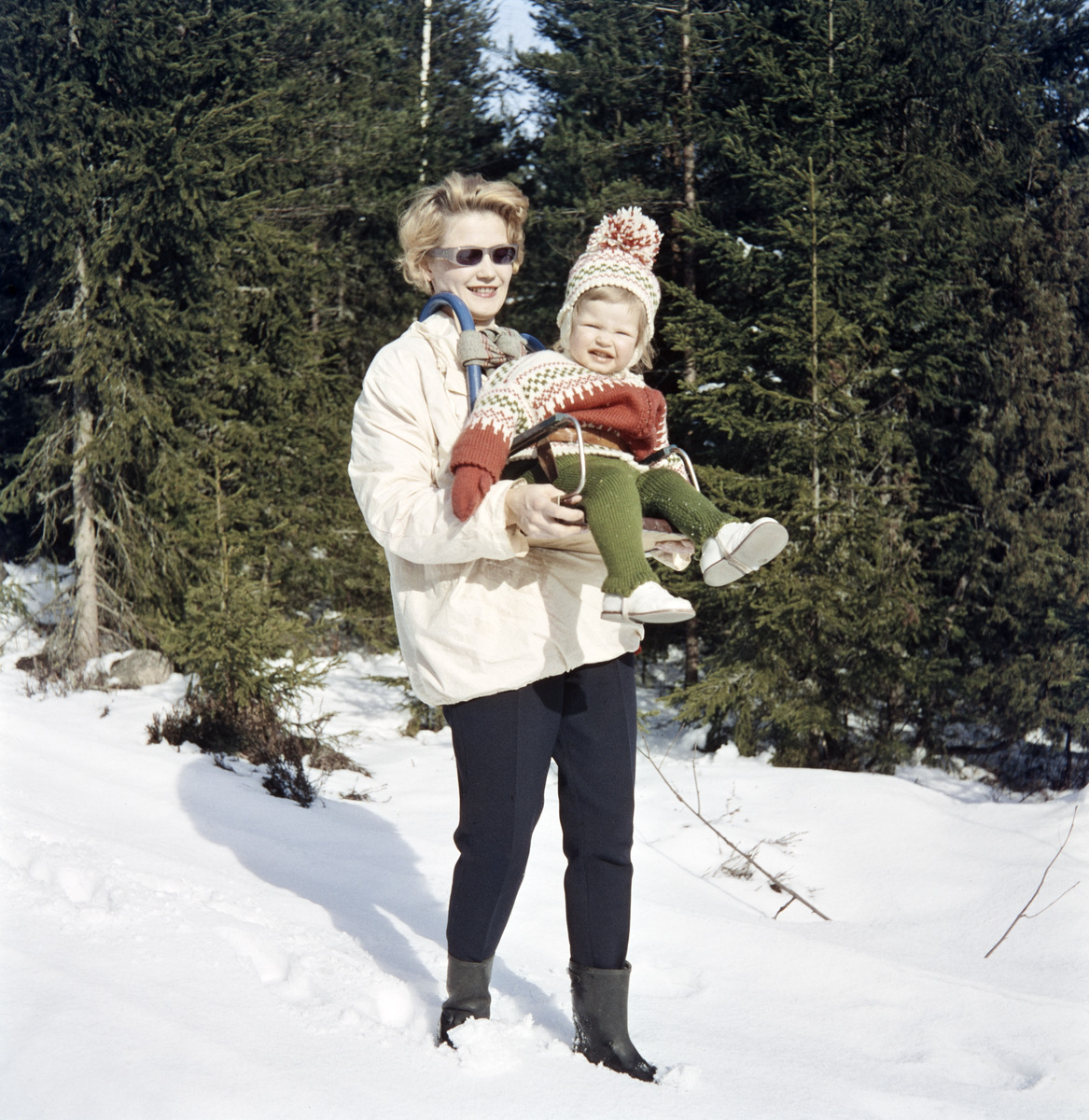 Äiti poseeraa kameralle hiihtoasussa tyttären kanssa metsän reunassa matkalla mökille Karjalohjalla