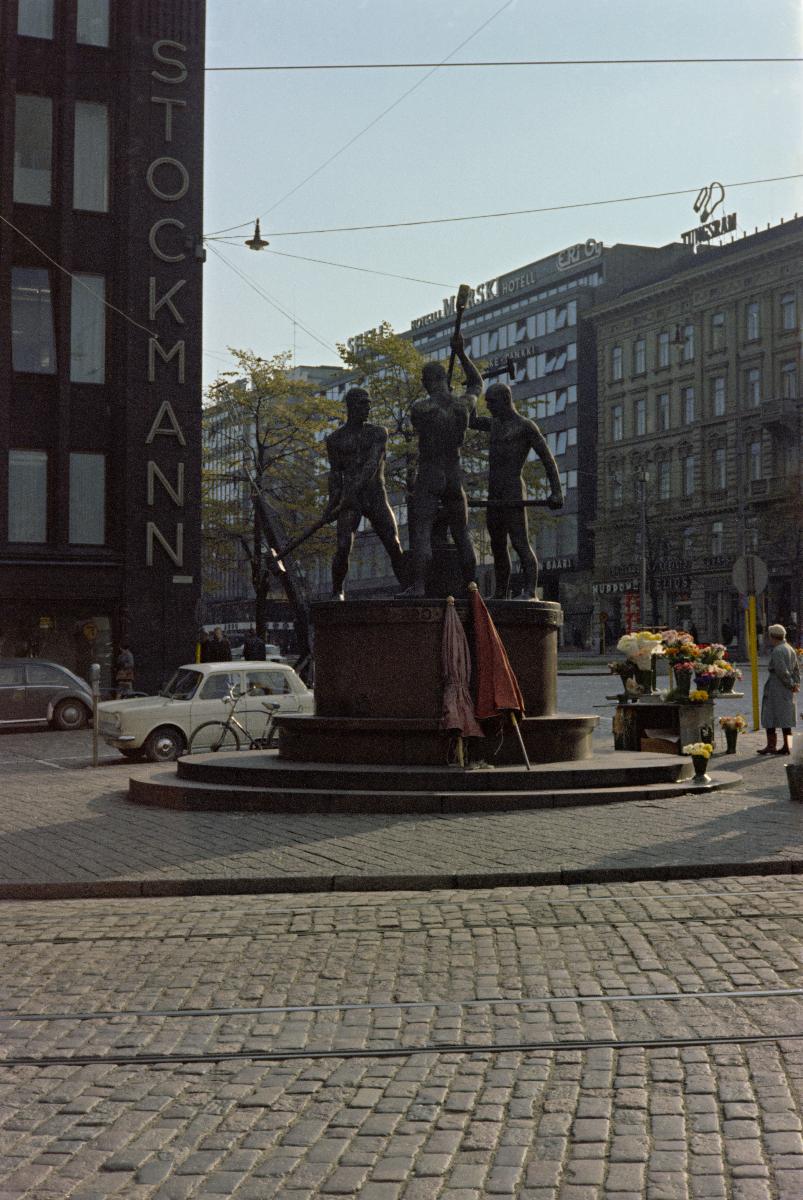 Kukkien myyntitiski ja kaksi kokoon taitettua päivänvarjoa Kolmen sepän patsaan (kuvanveistäjä Felix Nylund 1932) juurella.