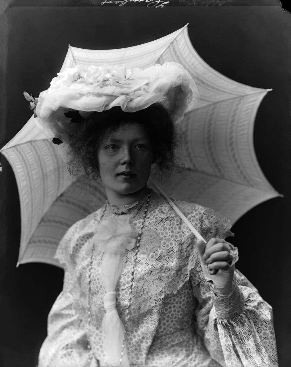 Oopperalaulaja Alfhild Hamberg (1881-1916) vaaleassa pitsikoristeisessa puvussa ja hatussa, kädessään päivänvarjo, joka nojaa olkapäähän