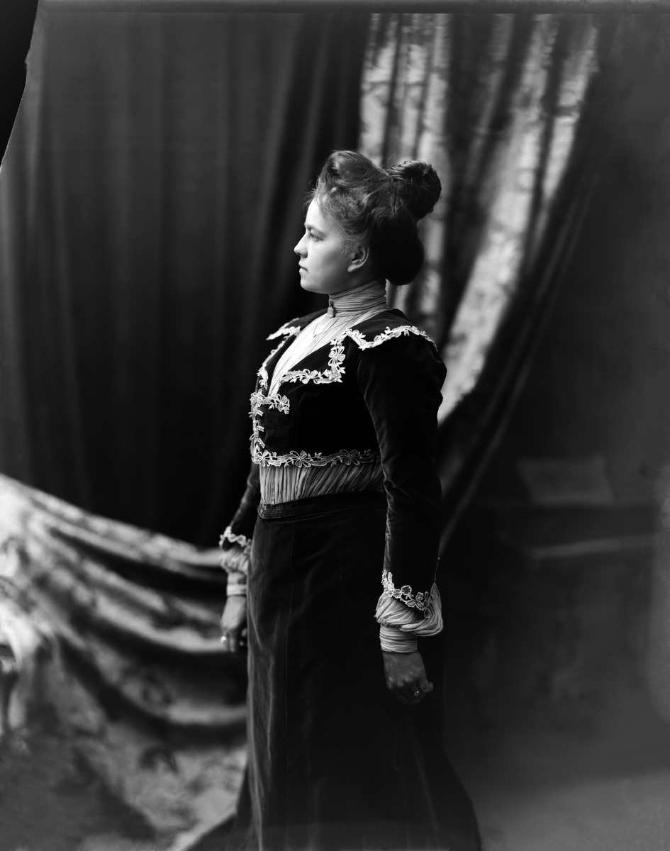 Nainen kuvattuna sivusta, yllään mekko jossa kukka-aihein kirjaillut kaulukset ja hihansuut