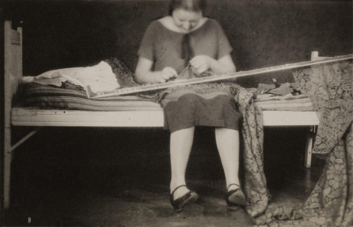Nainen, todennäköisesti keraamikko Friedl Holzer-Kjellberg, istumassa sängyllä, mahdollisesti kiinnittämässä verhoja verhojen pidikkeeseen tai kannattimeen